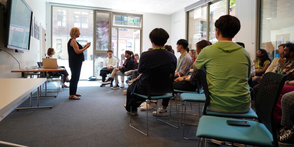 Students sit in a seminar room watching a speaker present at the front of the class.