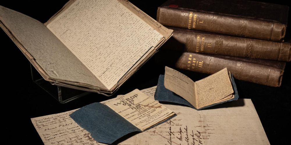 Two little books bound in paper originally used for Epsom Salts sit on top of a letter. Behind them, on the left is a handwritten book and on the right is a stack of first editions.