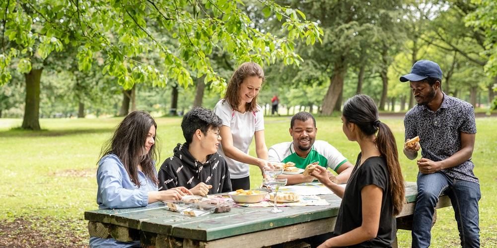 A group of students eating food sat on a picnic bench in a leafy park.