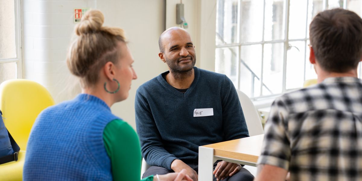 three individuals in conversation, the room is bright and light, they are wearing summery clothes.