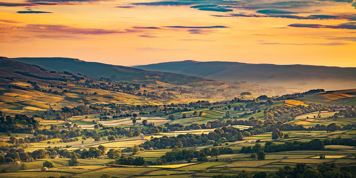 A valley in the Yorkshire Dales showing glowing sky, fields and rolling hills.