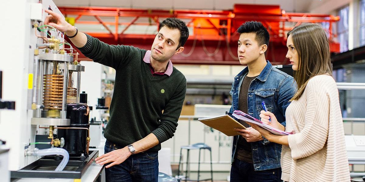 Two Mechanical Engineering students in a lab making notes as a lecturer points to a machine