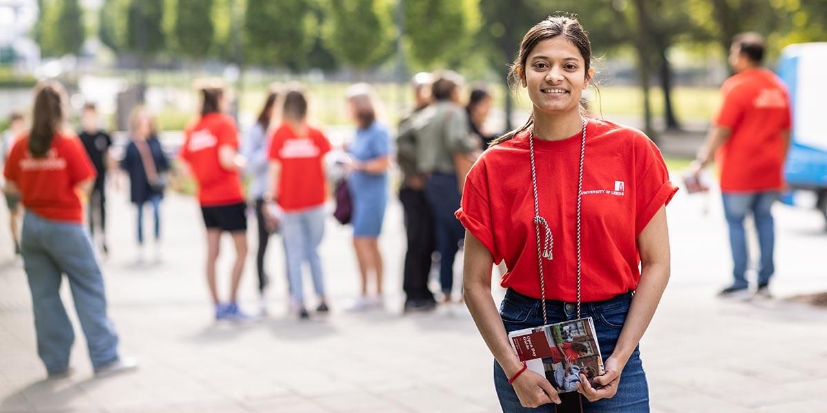 A Leeds student ambassador standing on campus, smiling, holding a brochure.