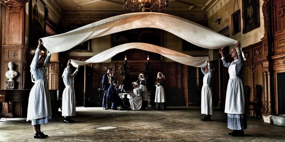 Performers dressed in historical costumes, as housemaids. They are lifting sheets above their heads, with other people in the background, in a historical looking room with wooden paneling on the walls