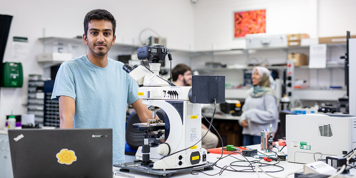 A male student standing in a lab behind a bench with his laptop open next to a microscope.