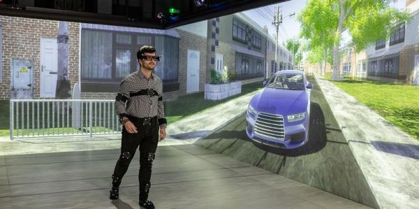 A man in striped with wearing monitoring equipment. Behind him are two screens that make up the pedestrian simulator, tne screens show a blue car approaching.