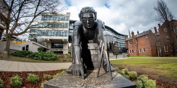 The bronze sculpture faces the camera head-on with campus in the background. The figure in the sculpture crouches over a compass. Its body is welded together, giving it a mechanical feel.