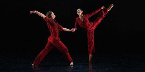 Two ballet dancers dressed in red pose against a black background, one in an arabesque