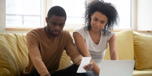 A couple sit on a sofa looking concerned as they read their bills.