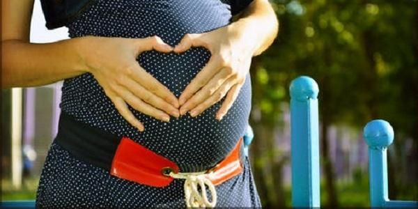 Picture shows a woman holding her pregnant tummy and making a heart shape with her hands