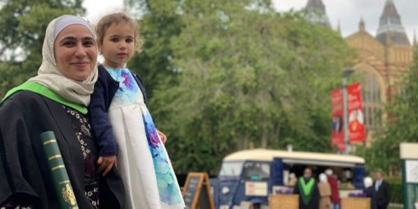 Loubna El Saboni in a graduation gown, with her young daughter, on campus. She is smiling and holding her degree.