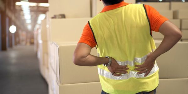 A male manual worker standing in front of some boxes in a warehouse and holding his back