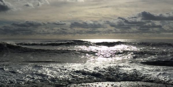 Seashore looking out to sea, gentle waves are coming into shore. There are clouds in the distance and the sun is hitting the water to create a glare.