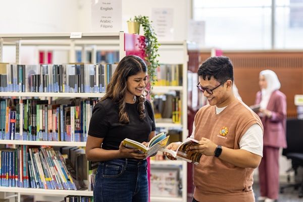 Two students in a library, looking at a book together. Book shelves are in the background.