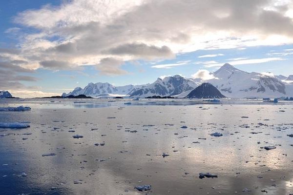 Image across a calm sea with floating ice debris to a coastline of ice and snow covered mountains. This is a the Antarctic Peninsula.