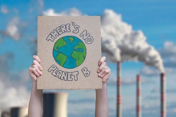 A pair of arms holding up a homemade sign that says "There's no planet B" with a power station in the background, pouring smoke out of its chimneys