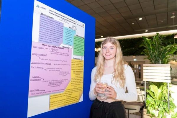 Kimberley Guy, smiling and holding a drink, standing next to a research poster.