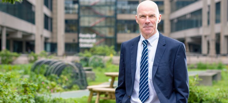 Mark Kearney, wearing a suit, standing in front of a University building with a garden and seating area behind him.