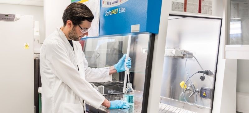 Dr Oliver Moore stands in a laboratory with a white lab tech coat on. He is standing at a counter and using a eye dropper to remove water from a clear bottle. 