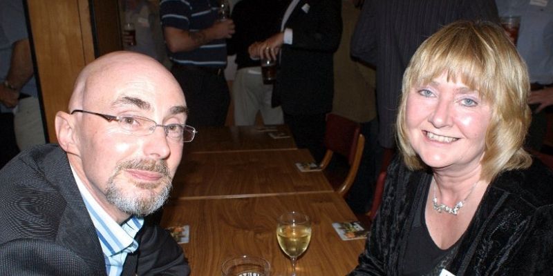 A couple smile at camera, wearing smart clothing and sitting in a bar