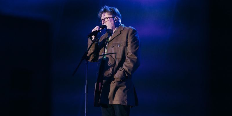 Professor Simon Armitage performs at the LEEDS 2023 Awakening event. He appears on a dark stage with a microphone, lit by a blue spotlight.