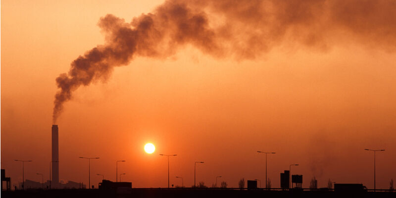 Smoke pouring out of a tall chimney in front of the setting orange sun.