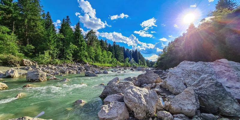 a river in full flow in the Alps, cascading over rocks in the foreground. In the background are trees and bright blue sky