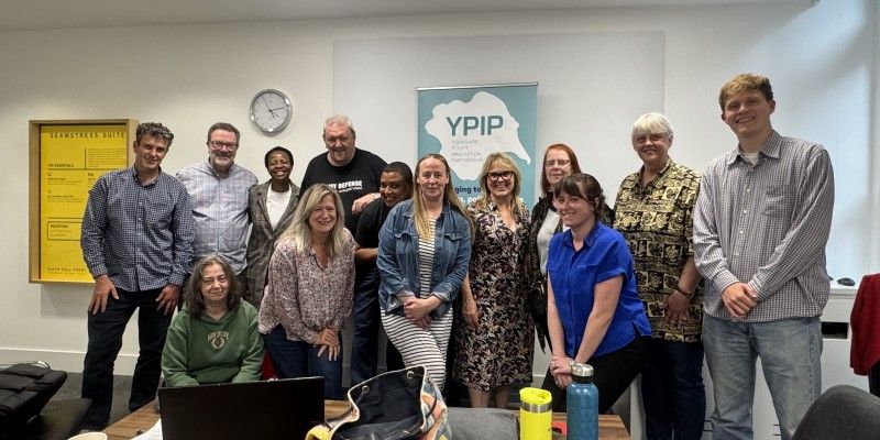 A group of 13 people stand in a conference room, in front of a sign saying "YPIP"