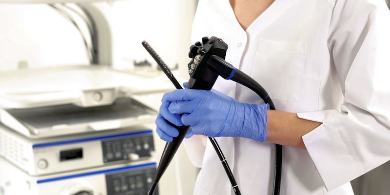 A female health professional in a white coat and blue gloves holds a colonoscopy device.