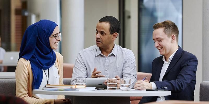 Three people in formal wear sat at a table. One holds a tablet and another is writing.