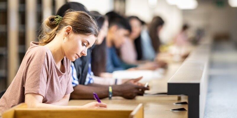 A student writing at a desk in the library, with other students working in the background.