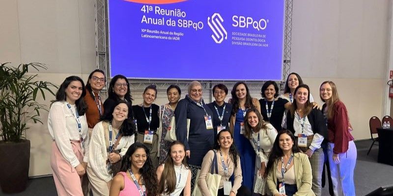 A large group of people stand smiling in front of a large screen with text in Spanish. They are smiling and wearing lanyards.