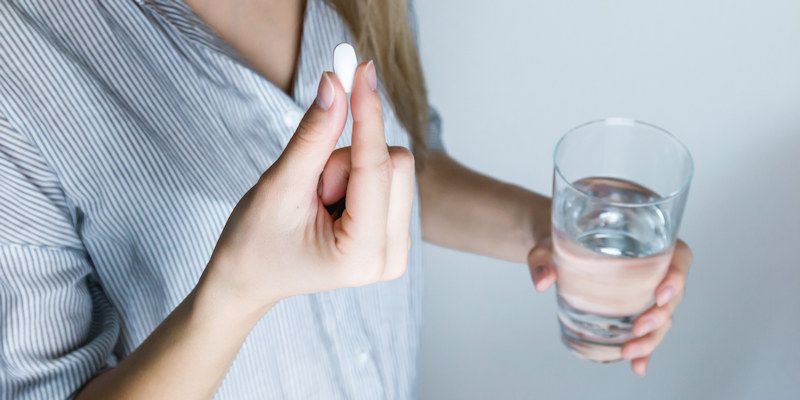 A woman in a nighty holds a white pill in her hand and a glass of water in the other
