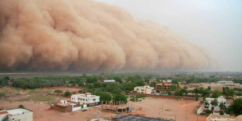 Overhead view of a village amongst green trees, with a large red dust cloud heading from the left over towards the houses.