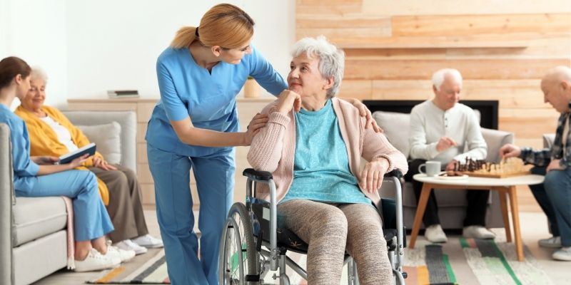 Older people in a care home setting. Two are playing chess and one person is looking at a book with a carer. Another person is sitting in a wheelchair talking to a carer.