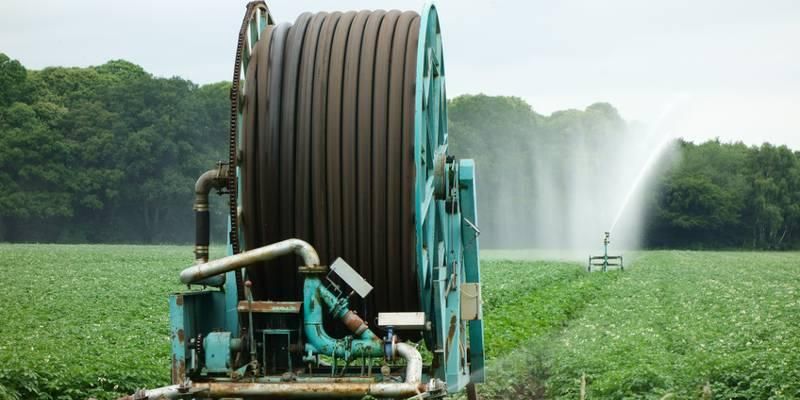 A spool with metres of hose wrapped around it stands on an open green field as water is sprayed out of a sprinkler in the distance.