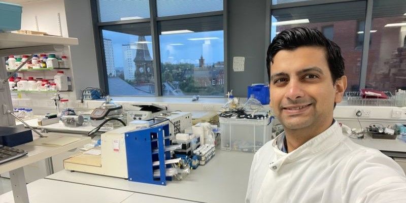 Omar stands taking a picture of himself ni the lab. He is smiling to the right of the picture in a lab coat and behind him is his laboratory bench and equipment.