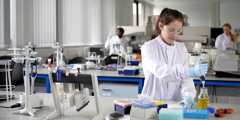 A young woman in a lab coat is holding a pipette and concentrating on moving liquids for a scientific experiment.