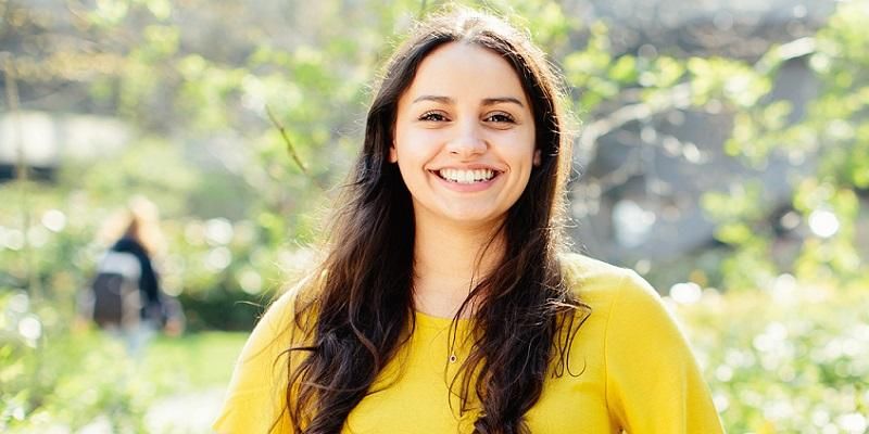 A Masters student smiling with trees and plants in the background on a sunny day.