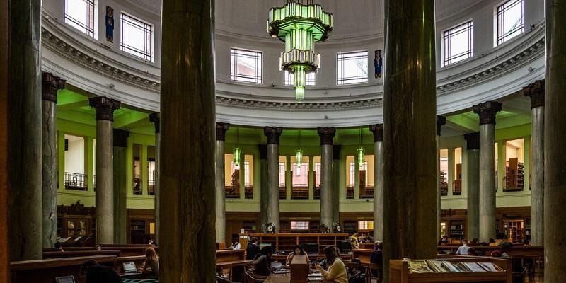 The image shows the grand interior of the rotunda of the Brotherton Library. An art deco light fitting hands from the ceiling. Students are sat at wooden desks studying.