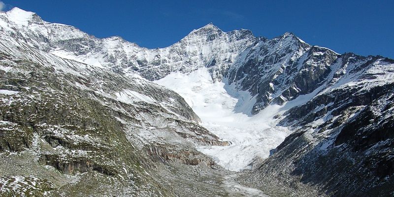 Odenwinkelkees glacier 
 with bare rocky mountainside on either side with bright blue sky in the background. 