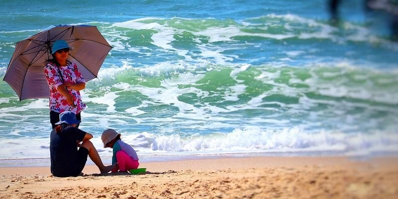 The image shows a family on a beach protected from the sun with an umbrella and wearing shorts and shirts