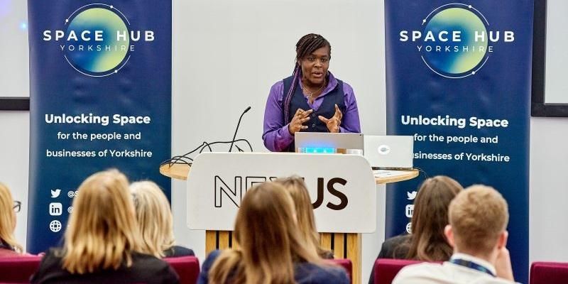 Dame Maggie Aderin-Pocock is at a lectern giving a lecture. From a hand gesture, it suggests she is explaining a complex concept.