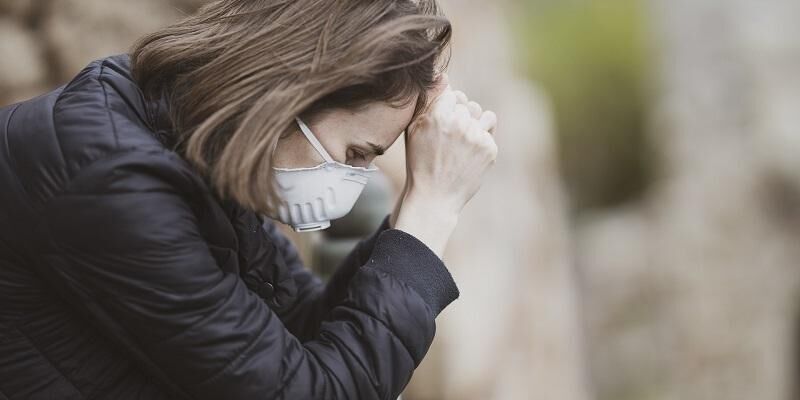 The image shows a woman, head in hands, wearing a face mask