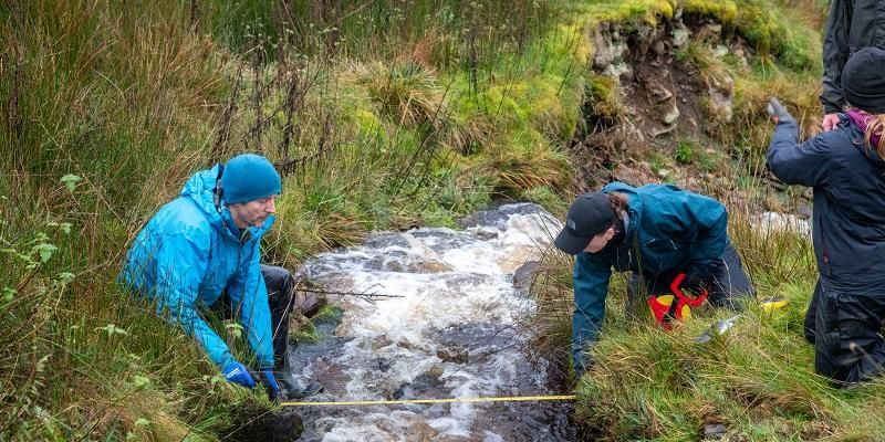 Two of the researchers are measuring a small but fast flowing stream. They are standing either side of the stream.