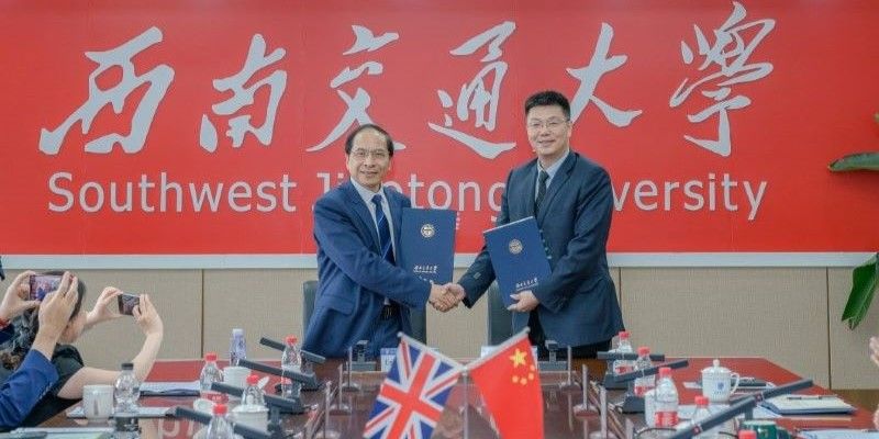 Deputy Vice-Chancellor Hai-Sui Yu stands shaking the hand of X at a conference table. The Union Jack and Chinese flag stand on the table, and behind them is a banner that says 'Southwest Jiaotong University'.