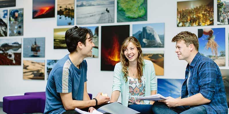 Students chatting and laughing together in a communal room on campus with posters on the wall behind them.