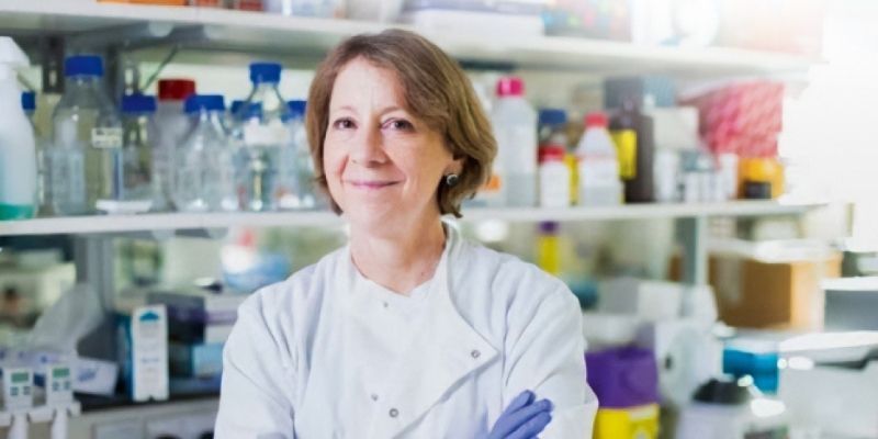 Professor Susan Short at camera in a lab coat with blue gloves on, with laboratory equipment behind her