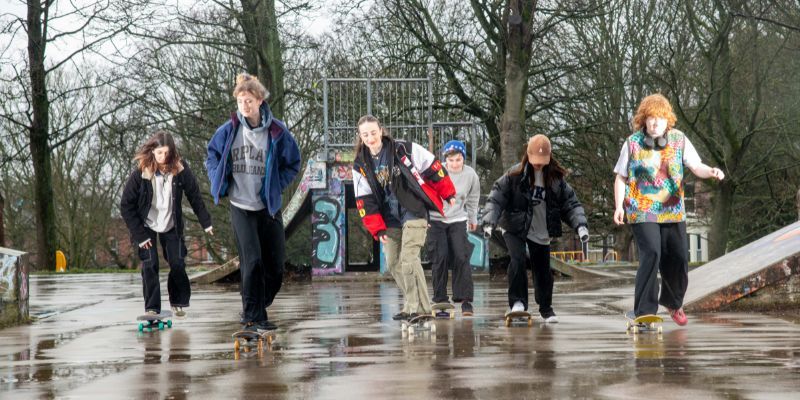 Six young women on skateboards skate towards the camera, laughing and smiling