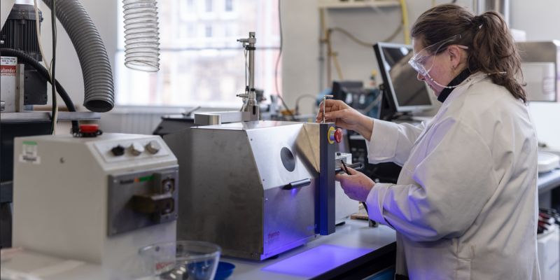 A lab technician in a white lab coat and safety goggles using a melt processing machine that emits a blue light.